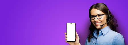 Customer support phone operator in glasses and headset holding showing smartphone cellphone mobile with white mock up screen on purple background. Consulting and assistance service call center.の写真素材