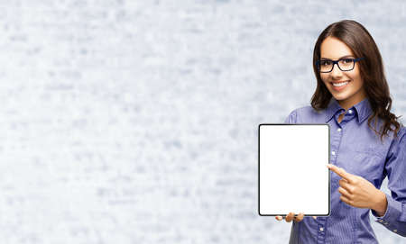 Happy smiling businesswoman in eye glasses spectacles showing touchpad tablet pc, with white screen copy space text area for advertising or slogan sign, over white bricks wall.の写真素材