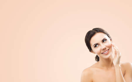 Portrait of beautiful dark haired woman touching, cleaning face skin, applying cream, over beige background. Amazed smiling brunette girl at studio. Beauty model picture. Copy space empty text area.の写真素材