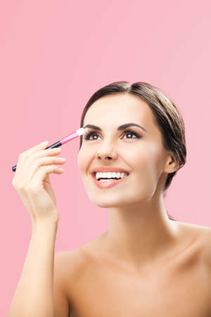 Portrait of beautiful dark haired woman with makeup cosmetics brush, isolated over rose pink background. Happy smiling brunette girl looking up, at studio. Beauty model image.の写真素材