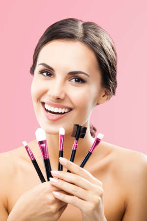 Portrait of beautiful dark haired woman holding in hands, showing, giving cosmetics brushes, isolated over rose pink background. Happy smiling brunette girl at studio. Beauty treatment model picture.の写真素材