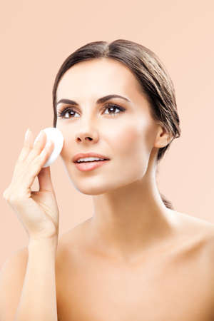 Portrait of beautiful dark haired woman cleaning face skin by cotton pad, isolated over beige background. Happy smiling brunette girl looking up, at studio. Beauty model picture.の写真素材