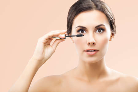Portrait of beautiful woman holding in hand, putting mascara on long thick eye lashes with brush, isolated over beige background. Brunette girl at beauty treatment, visage, makeup studio concept.の写真素材