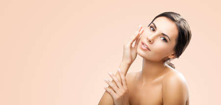 Portrait of beautiful dark haired woman touching her perfect face skin or applying cream, isolated over beige background. Attractive brunette girl at studio. Beauty salon, cosmetics concept image.の写真素材