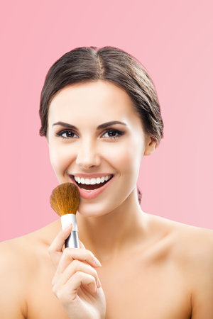 Portrait of beautiful excited smiling woman holding in hand, showing, giving cosmetics brush, isolated over rose pink color background. Happy brunette girl at beauty treatment, visage, makeup concept.の写真素材