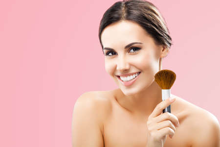 Portrait of beautiful smiling woman holding in hand, showing, giving cosmetics brush, isolated on rose pink background. Happy amazed brunette girl at beauty treatment, visage, makeup studio concept.の写真素材