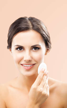 Portrait of beautiful dark haired woman cleaning face skin by cotton pad, isolated over beige background. Happy smiling brunette girl at studio. Beauty model picture.の写真素材