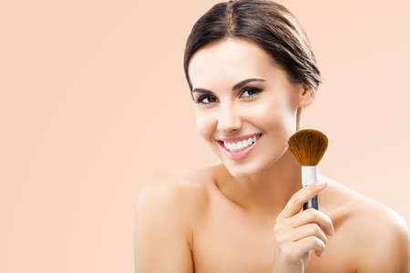 Portrait of beautiful smiling woman holding in hand, showing, giving cosmetics brush, isolated over beige background. Happy amazed brunette girl at beauty treatment, visage, makeup studio conceptの写真素材