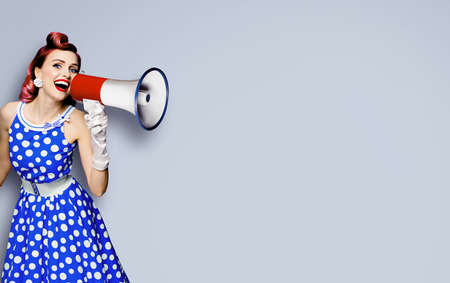 Portrait of purple haired woman holding mega phone, shout advertising something. Girl in blue pin up style dress with mega phone loudspeaker. Isolated on grey background. Beauty model in retro conceptの写真素材