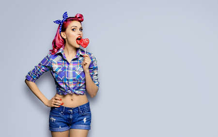 Portrait of beautiful woman licking heart shape lollipop dressed in pinup plaid shirt, isolated over grey background. Purple red girl in retro fashion and vintage studio concept. Mock up.の写真素材