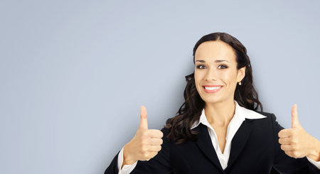 Businesswoman in black confident suit showing thumbs up gesture, over grey color background. Happy smiling gesturing brunette woman at studio. Business concept photo.の写真素材