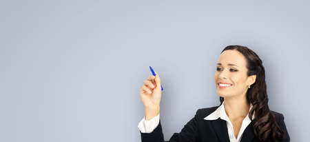 Portrait of happy smiling beautiful young businesswoman in black confident suit, writing or drawing something on screen, by marker. Brunette woman, isolated over grey color backgroundの写真素材
