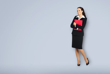 Full body length of happy smiling businesswoman holding red folder, copy space area for text. Confident woman in black suit standing over grey background. Business concept.の写真素材