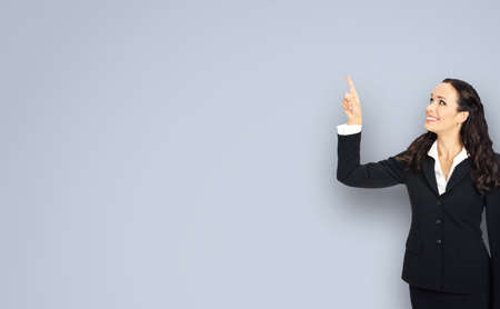Profile portrait of smiling executive woman in black confident suit, pointing showing up copy space. Business concept. Grey background. Brunette businesswoman.の写真素材