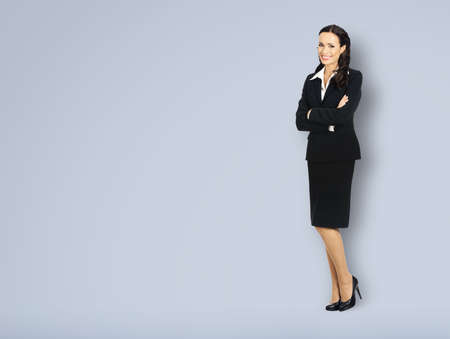 Full body length of happy smiling businesswoman with crossed arms, with copy space area for some text. Confident woman in black suit standing on grey background. Business concept.の写真素材