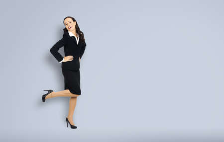 Full body length of happy excited smiling businesswoman with hands on hips, copy space for text. Confident woman in black suit standing dancing on grey background. Business concept.の写真素材