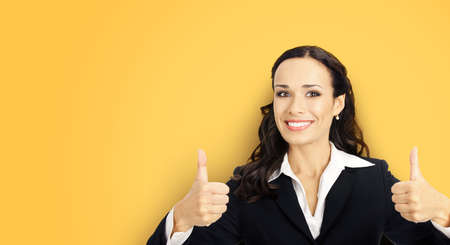 Businesswoman in black confident suit showing thumbs up gesture, yellow color background. Happy smiling gesturing brunette woman at studio. Business concept photo.の写真素材