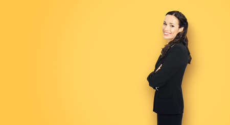 Happy smiling attractive businesswoman, with copy space area for some slogan or advertising text. Confident brunette woman in black suit standing on yellow color background. Business ad concept.の写真素材