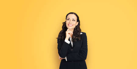 Happy smiling amazed positive businesswoman looking up, with copy space for slogan or text. Confident brunette business woman in black suit, on yellow color background.の写真素材