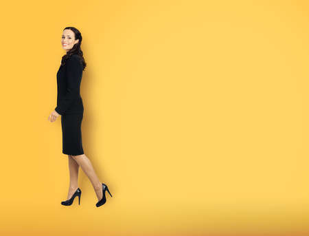 Full body length profile side photo of happy excited smiling businesswoman going side away, over copy space area. Waking confident business woman in black suit, yellow color background.の写真素材