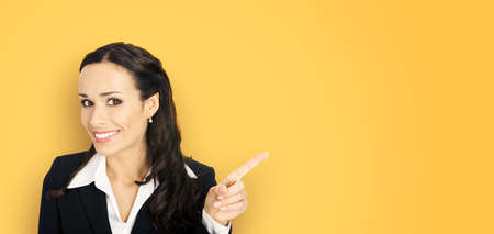 Young attractive confident woman in black suit show pointing finger at something or copy space empty text offer area. Business ad concept. Yellow color background. Brunette businesswoman. Wide image.の写真素材