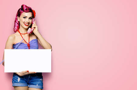 Woman with phone, in pinup cloth, showing blank white signboard with mock up copy space free text area, pink background. Red purple hair girl pose in retro fashion vintage studio concept.の写真素材