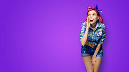 Happy excited woman holds hand near open mouth, looking aside and saying something. Girl dressed in pinup style. Red purple haired pin up model at retro fashion vintage concept, violet background.の写真素材