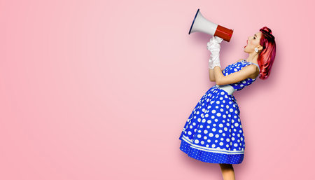 Red purple haired woman holding red megaphone, shout advertising something. Girl in blue pin up style dress in polka dot. Rose pink background with mock up. Female model in retro vintage studio photoの写真素材
