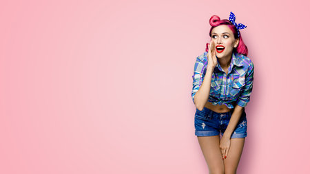 Happy excited woman holds hand near open mouth, looking aside and saying something. Girl dressed in pinup style. Red purple haired pin up model at retro fashion vintage concept, pink background.の写真素材