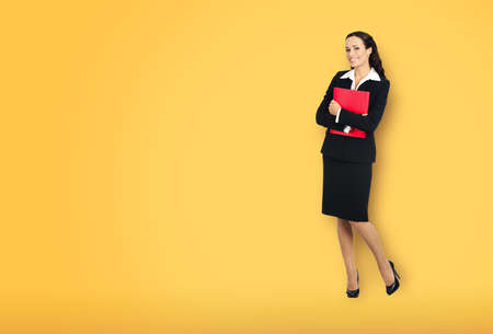 Full body length of happy smiling businesswoman holding red folder, copy space area for text. Confident business woman in black suit standing over yellow color background.の写真素材