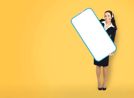 Call Center Service. Customer support, sales agent. Businesswoman in headset or phone operator hold show cellphone mobile white screen. Full body business woman with big smartphone. Yellow background.の写真素材