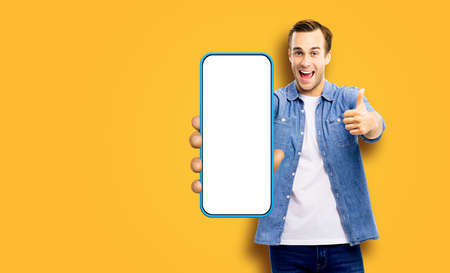 WOW! Great mobile app. Excited young man, showing cell phone, mobile smartphone, Happy guy hold cellphone with white screen, isolated over orange yellow studio wall background. Advertisement ad offer.の写真素材