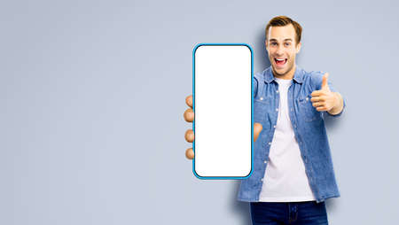 WOW! Great mobile app. Excited young man, showing cell phone, mobile smartphone, Happy guy hold cellphone with white screen, isolated over grey gray studio wall background. Advertisement ad offer.の写真素材
