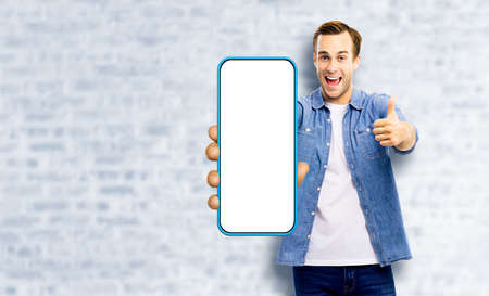 WOW! Great mobile app. Excited young man, showing cell phone, mobile smartphone, Happy guy hold cellphone with white screen, standing over loft style wall background. Advertisement ad offer.の写真素材