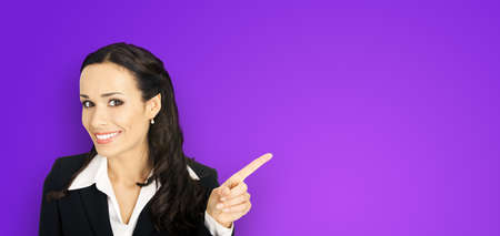 Young attractive confident woman in black suit show pointing finger at something or copy space empty text area. Business ad concept. Violet purple background. Brunette businesswoman. Wide imageの写真素材