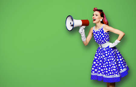 Purple haired woman holding red megaphone, shout advertising something. Girl in blue pin up style with mega phone loudspeaker. Green background with mock up. Female model in retro fashion dress.の写真素材