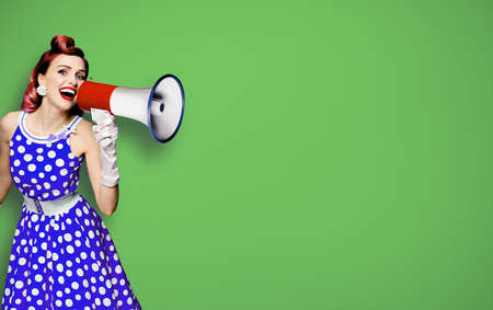 Portrait image of purple haired woman holding mega phone, shout advertising something. Girl in blue pin up style dress with mega phone loudspeaker. Isolated on green background. Beauty model.の写真素材