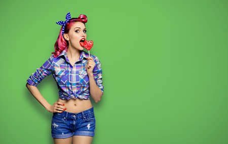Portrait of beautiful woman licking heart shape lollipop dressed in pinup plaid shirt, isolated over green background. Purple red girl in retro fashion and vintage studio concept.の写真素材