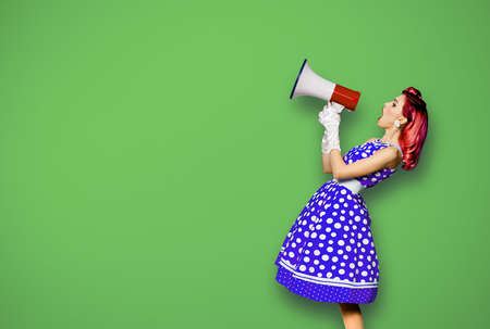 Red purple haired woman holding red megaphone, shout advertising something. Girl in blue pin up style dress in polka dot. Green background with mock up. Female model in retro vintage studio photoの写真素材