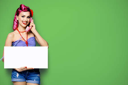 Woman with phone, in pinup cloth, showing blank white signboard with mock up copy space free text area, green background. Red purple hair girl pose in retro vintage studio concept.の写真素材