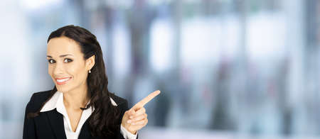 Young attractive confident woman in black suit show pointing finger at something or copy space empty text area. Business ad concept. Blurred office background. Brunette businesswoman. Wide imageの写真素材