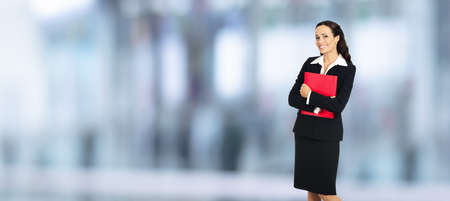 Portrait of happy smiling businesswoman holding red folder, copy space area for text. Confident business woman in black suit standing over blurred office background.の写真素材