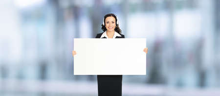 Call Center Service. Customer support or sales agent. Businesswoman or caller phone operator hold white banner. Business woman in headset with empty signboard. Blurred office background.の写真素材