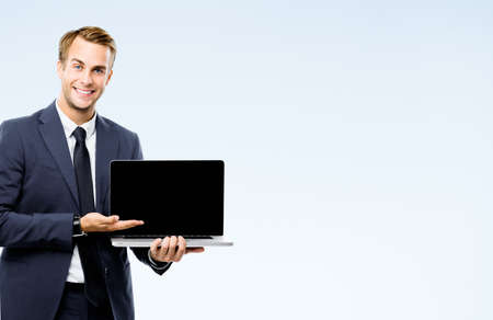 Happy smiling young business man in black suit, necktie showing blank laptop monitor screen, isolated on grey background, with copy space area. Businessman holding notebook computer at studio image.の写真素材