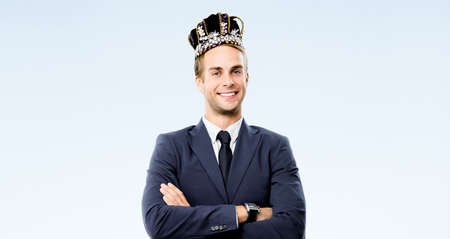 Portrait image of excited happy smiling business man in fake crown on his head, crossed arms pose, isolated over grey background. Young businessman at leadership success studio ad concept.の写真素材
