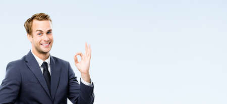 Happy smiling businessman in black suit, white shirt, tie showing okay hand sign gesture, isolated on grey background. Young business man at studio image. Success ad concept. Copy spaceの写真素材