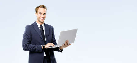 Happy smiling young business man in black suit, necktie using laptop computer, isolated on grey background, with copy space area for ad text. Businessman holding notebook at studio concept image.の写真素材