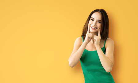 Bright image of excited amazed brunette woman showing pointing finger her smile, green casual cloth, isolated on yellow color background. Optimistic, positive, happy feeling or dental care ad concept.の写真素材