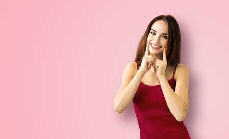 Bright image of excited amazed brunette woman showing pointing finger her smile, in red clothing, isolated over rose pink background. Optimistic, positive happy feeling or dental care ad conceptの写真素材