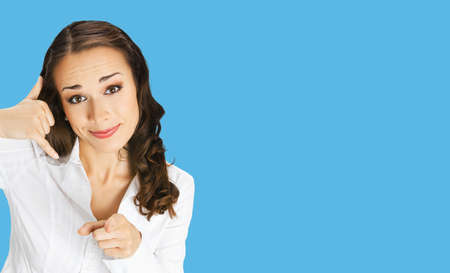 Businesswoman in white confident clothing showing call me hand sign gesture and pointing at viewer, isolated on blue background. Portrait of smiling brunette woman at studio. Business concept.の写真素材